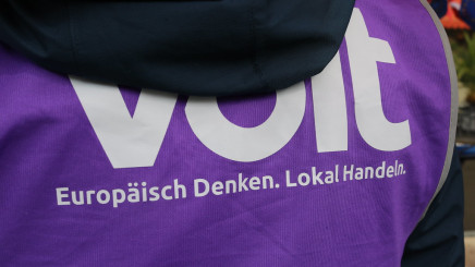 Nahaufnahme des Rückens einer Person, die über einer dunkelblauen Jacke eine leuchtend violette Kampagnenweste trägt. Auf der Weste ist in großen weißen Buchstaben der Schriftzug „Volt“ aufgedruckt, der oben teilweise von der Kapuze verdeckt wird, darunter der weiße Slogan: „Europäisch Denken. Lokal Handeln.“