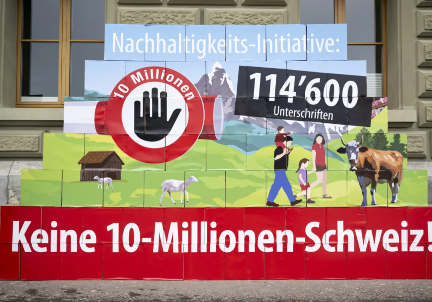 Eine Pyramide aus bedruckten Boxen vor einer Steinfassade zeigt eine ländliche Schweizer Landschaft mit Bergen, einer Familie, einer Kuh und Schafen. Darauf integriert sind ein Stoppsymbol mit einer Hand und dem Text «10 Millionen» sowie die Schriftzüge «Nachhaltigkeits-Initiative:», «114'600 Unterschriften» und gross auf rotem Grund am Fuss der Pyramide «Keine 10-Millionen-Schweiz!».