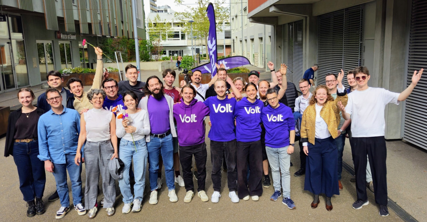 Eine Gruppe von etwa 20 gut gelaunten Menschen posiert im Freien auf einem asphaltierten Platz vor modernen Gebäuden. Mehrere Personen in der Mitte tragen violette T-Shirts oder Hoodies mit dem weissen Aufdruck «Volt». Einige Personen halten Blumen hoch oder haben die Arme jubelnd in die Luft gestreckt. Im Hintergrund ist eine violette Volt-Fahne zu sehen. Das Bild vermittelt eine positive, gemeinschaftliche Atmosphäre.