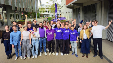 Eine Gruppe von etwa 20 gut gelaunten Menschen posiert im Freien auf einem asphaltierten Platz vor modernen Gebäuden. Mehrere Personen in der Mitte tragen violette T-Shirts oder Hoodies mit dem weissen Aufdruck «Volt». Einige Personen halten Blumen hoch oder haben die Arme jubelnd in die Luft gestreckt. Im Hintergrund ist eine violette Volt-Fahne zu sehen. Das Bild vermittelt eine positive, gemeinschaftliche Atmosphäre.